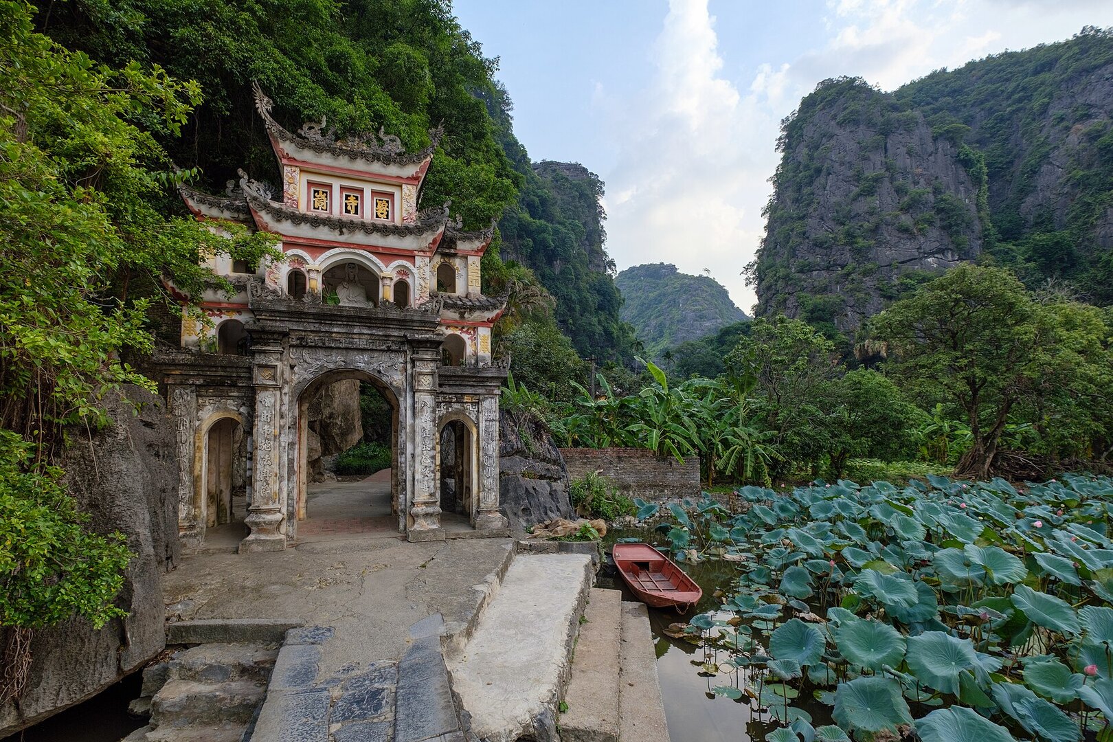 Ao sen trước cổng chùa Bích Động giữa núi đá vôi, lá sen xanh xen nụ và hoa sen hồng, một chiếc thuyền đỏ đậu ở bậc đá.