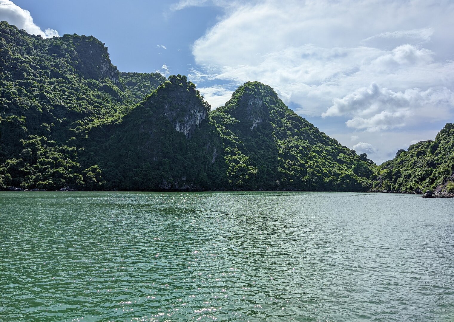 Đảo đá vôi phủ rừng xanh trên vịnh Hạ Long, nước ngọc lục bảo dưới trời nắng nhiều mây.