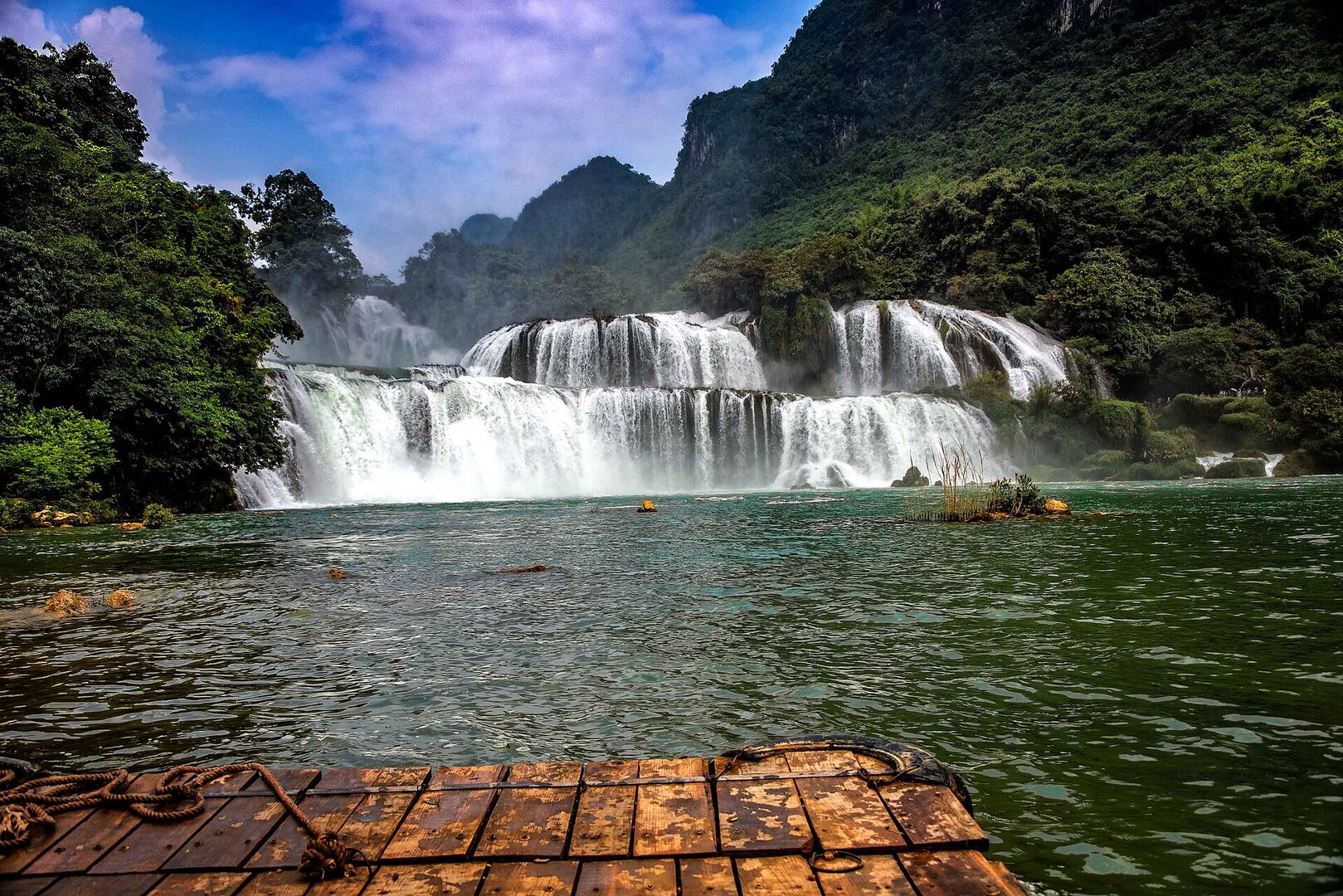 Ban Gioc Waterfall in full flow, white cascades pouring over several tiers into a green river, seen from a bamboo raft.
