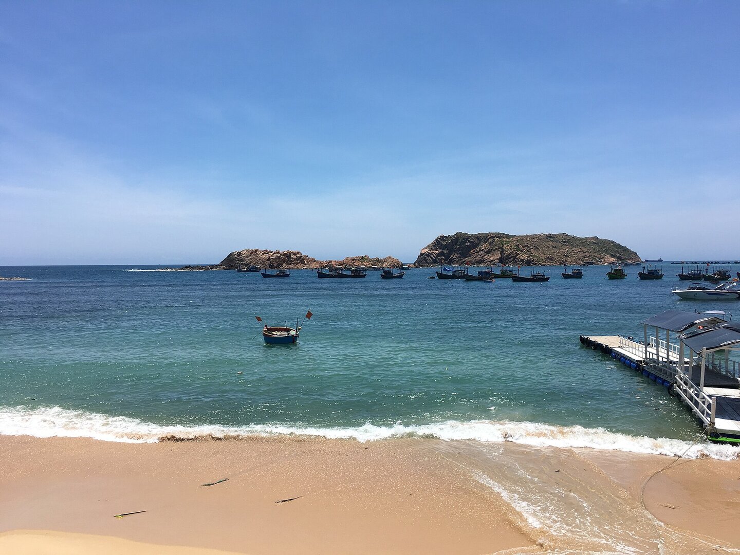 Biển Nhơn Hải (Quy Nhơn) trong xanh một trưa tháng Tư, thuyền thúng và tàu cá neo gần bờ, hòn đảo đá phía xa.